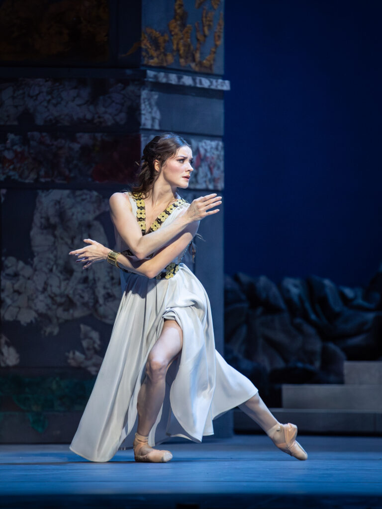 Trenary looks over her shoulder as she wraps her arms around herself, pausing in a low lunge. Her white, Grecian-style dress drapes around her legs to skim the floor.