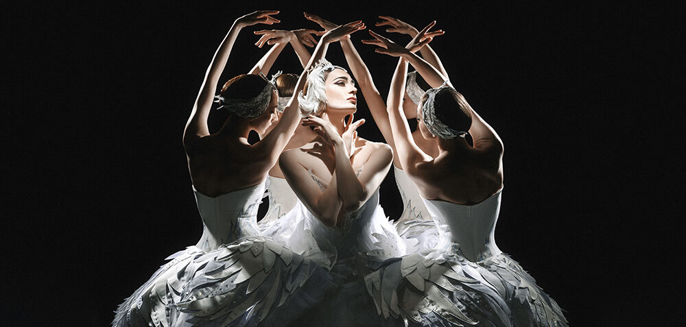 Lit overhead by a single spotlight against a pitch black background, a circle of ballet dancers dressed as swans surround one principal, dressed as Odette. The corps dancers lean slightly inward with arms overhead, fingertips brushing in, with their backs facing out. In the middle, Odette turns her face and crosses her arms so her fingertips brush her neck on either side.