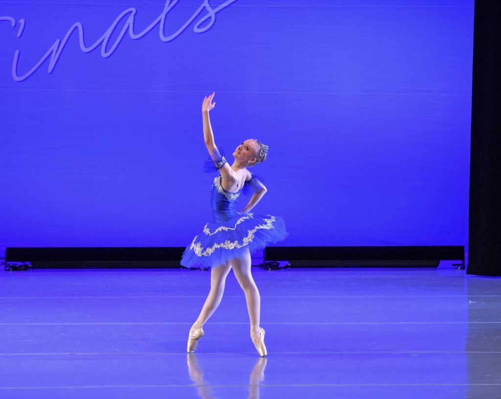 Dulcianna Jonak poses in fourth position on pointe with one hand on her hip and the other scooping up high. She twists her shoulders for epaulement and wears a blue tutu with white and silver accents.