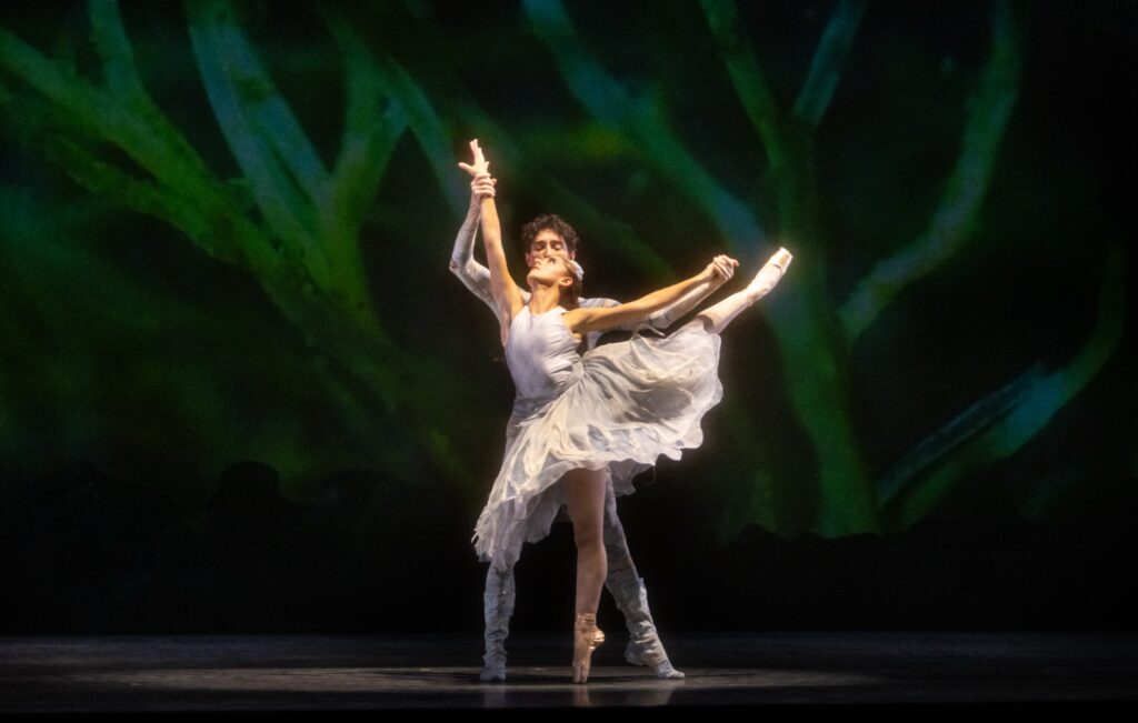 Wearing a grey dress, Taylor Naturkas poses en pointe in an arabesque with her right leg extended behind her. Damian Zamorano supports her by holding both of her hands. A green patterned backdrop fills the background.