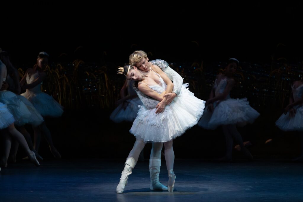 In a white tutu and a gold crown, Taylor Naturkas stands en pointe on her right leg with her left eelevated behind her. Brooks Landegger stands behind her and wraps his arms around her in an embrace.