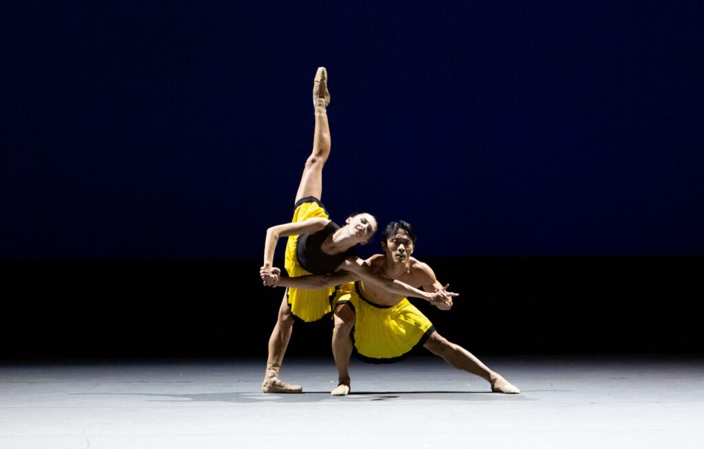 Two dancers appear on stage both wearing bright yellow skirts with a black trim at the bottom. Shimon Ito is in a side lunge as he holds his partners hands and looks out at the audience. Taylor Naturkas wears nude pointe shoes and poses in a penché next to Ito with their arms intertwined.