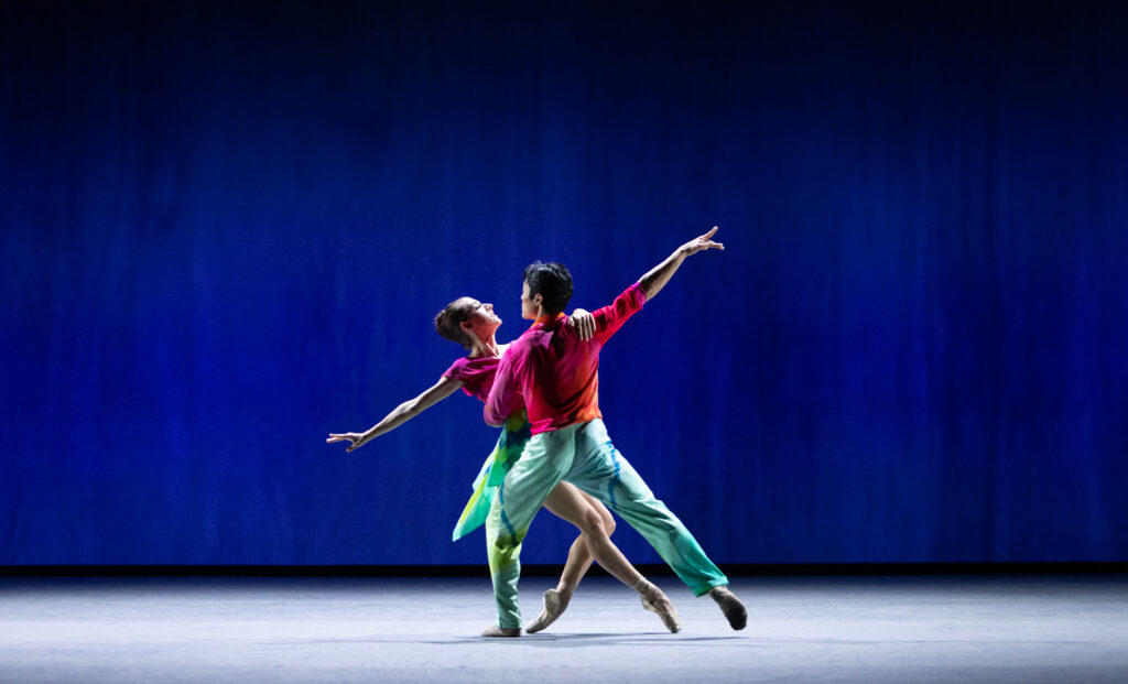 Shimon Ito lunges on his left leg facing upstage and holds Taylor Naturkas by the waist. Naturkas balances on pointe with her left leg bent and tucked underneath her and her right leg extended, and looks Ito as she leans back slightly in his arms. They wear pink and red tops, with Naturkas in a green skirt and Ito in matching pants.