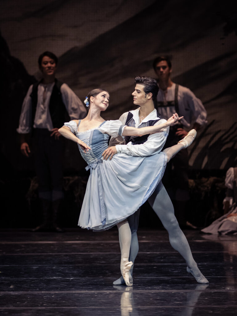 renary smiles back at her partner as he supports her at the waist in a back attitude balance en pointe. They are costumed like peasants.