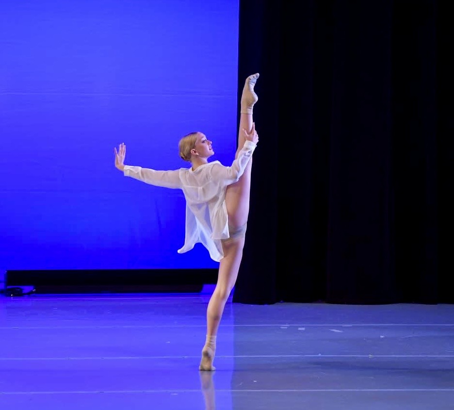 Kenedi Sampson, in a white button-down over a gray leotard, battements one leg on relevé and holds the working leg with one hand. Her other arm extends back with a flexed palm.