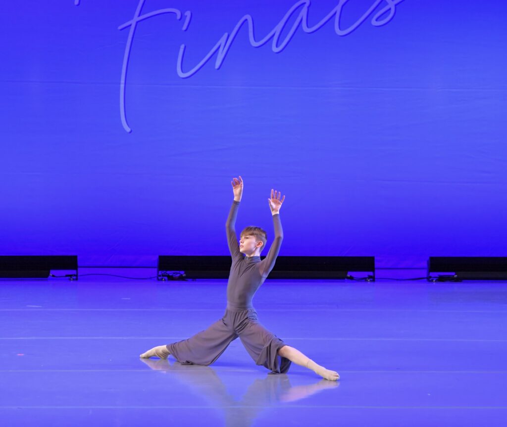 Hunter Moreland, in a long-sleeved gray costume with loose pant legs, lifts his arms up as he pushes himself up with his legs from a middle split.