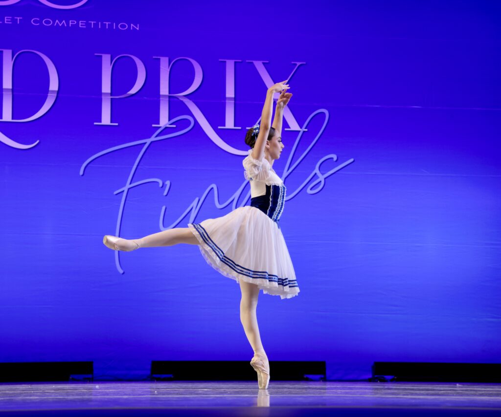 Lena Garcia does a piqué arabesque on pointe, facing the right, with her arms in high fifth. She wears a blue and white peasant costume.