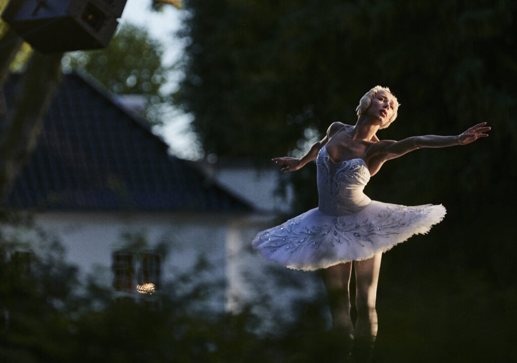 Wearing a white tutu and head piece, Iana Salenko extends both of her arms reaching. She appears outside with a building and trees behinid her.