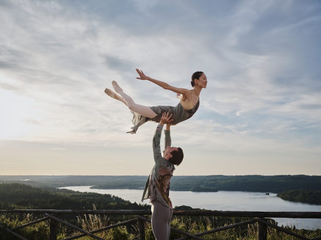 Against a backdrop of a river and lush green landscape, Gareth Haw angel lifts Sangeun Lee. Both dancers are dressed in gray.
