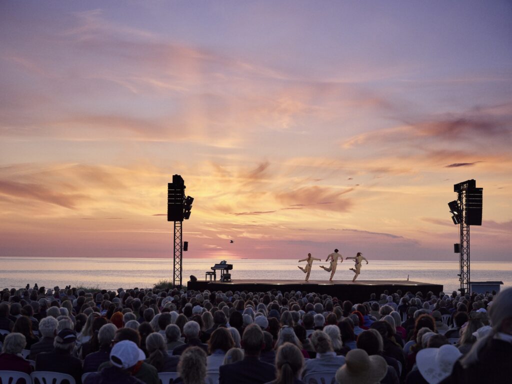Three dancers appear on an outdoor stage with a large body of water and a beautiful sunset in the background. They are all dressed in matching beige shirts and pants. Each dancer is in a turned-in passé position, with their knees touching.