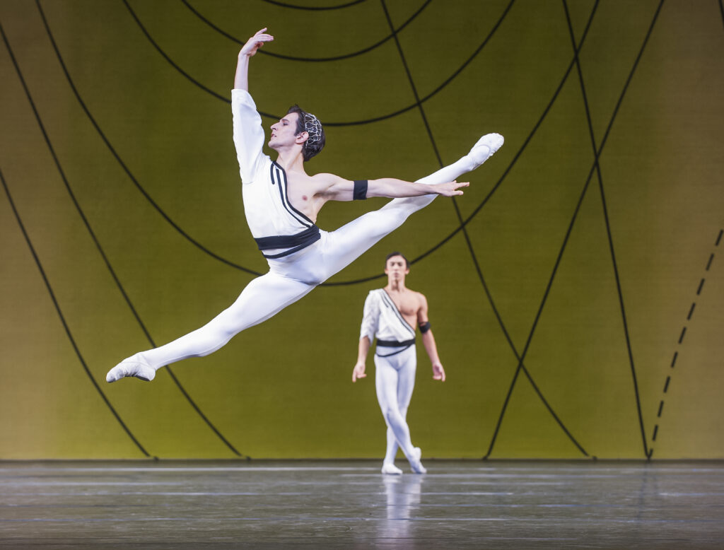 Valentino Zucchetti splits his legs in a powerful sauté, arms in high third. He wears white tights and a white one-sleeved tunic with black accents. Another dancer stands onstage behind him with one leg casually crossed over the other.