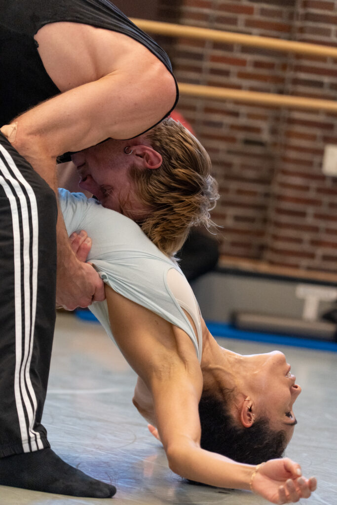Silas Henriksen and Nora Kimball rehearse a contemporary pas de deux in a studio with brick walls. Henrickson bends over with relaxed knees as he holds Kimball, who folds in half backwards, around her waist. He rests his head on her chest as she lets her arms hang loose overhead, resting gently on the floor.