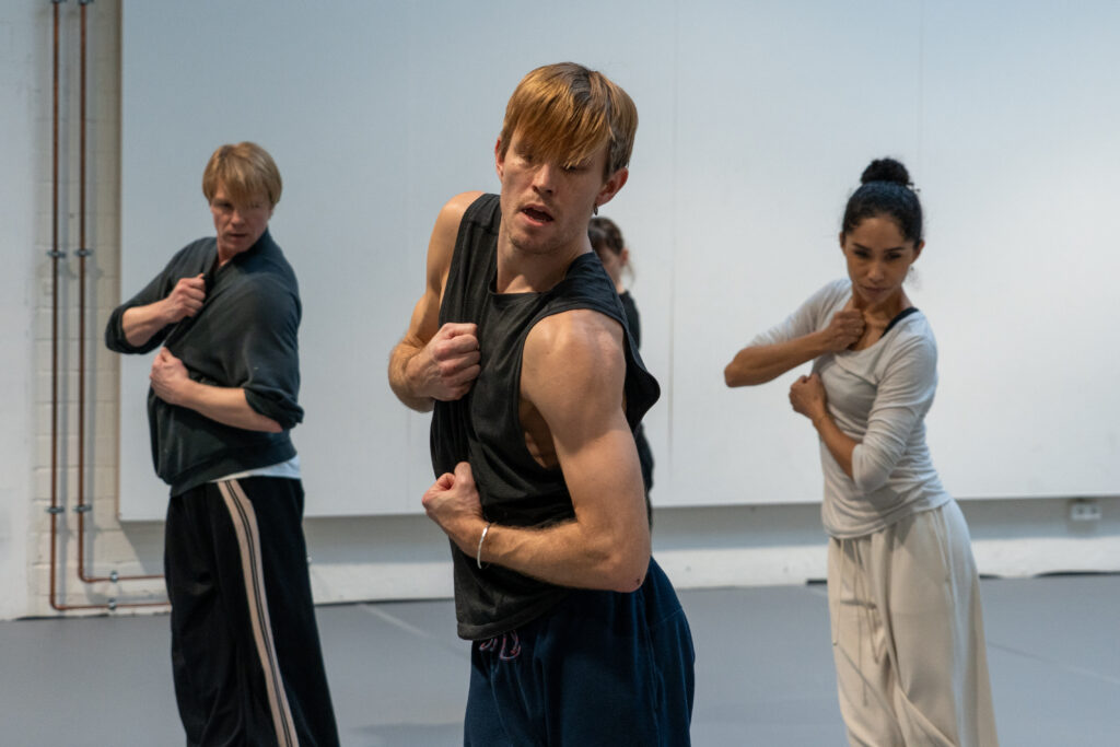 Four dancers in loose-fitting practice clothes stand in a diamond shape. They twist their torsos with their hips facing to the left as they tuck one arm in toward their chests and flex the other to the front, elbow bent.