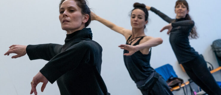 Igone de Jongh, Diana Vishneva, and Mara Galeazzi stand in a diagonal line with one arm lifted to the side and the other lifting gently front with the elbow bent. They wear dark-colored practice clothes.