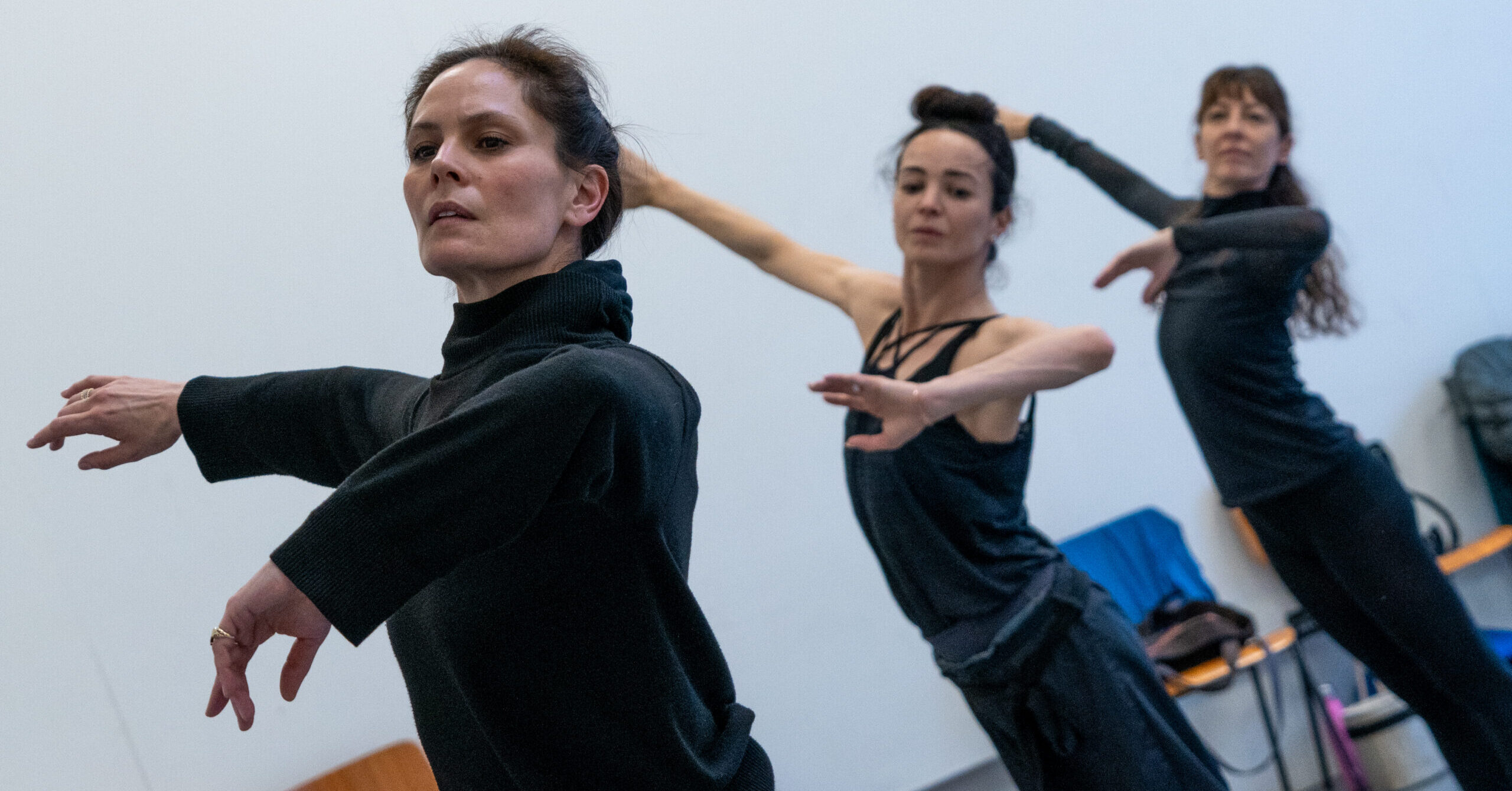 Igone de Jongh, Diana Vishneva, and Mara Galeazzi stand in a diagonal line with one arm lifted to the side and the other lifting gently front with the elbow bent. They wear dark-colored practice clothes.