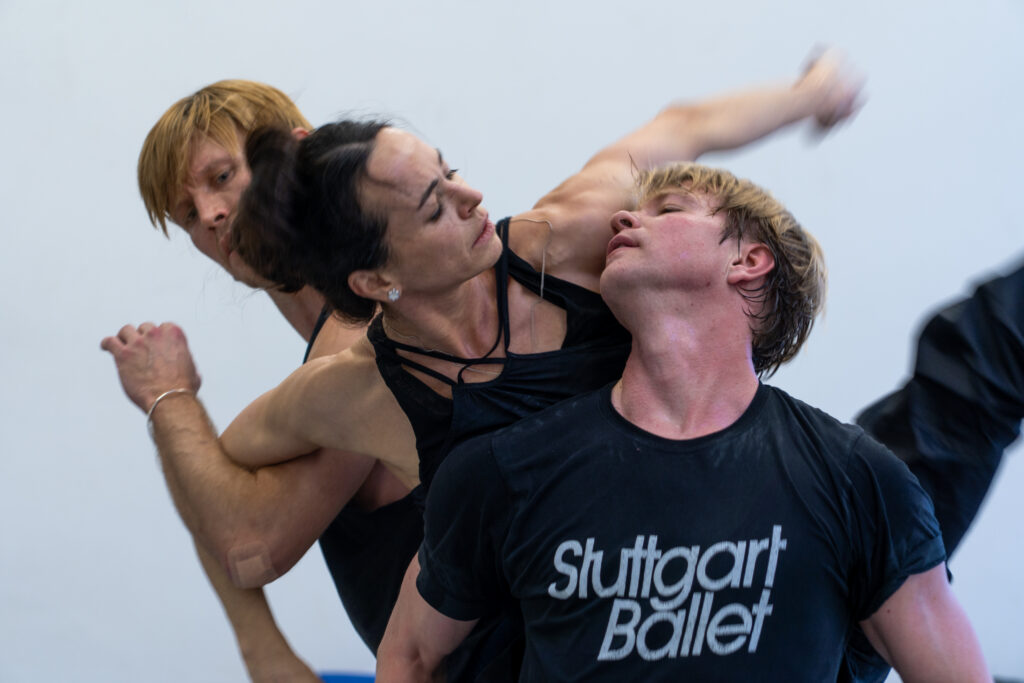 Silas Henriksen, Diana Vishneva, and Marijn Rademaker share their weight in rehearsal wearing dark-colored practice clothes. Vishneva bends forward slightly with her arms extended out and down, looking into Rademaker's eyes as she leans on his right shoulder. Henriksen pumps one arm while turning his head toward them. He and Vishneva link one arm as she lifts one leg to the side.