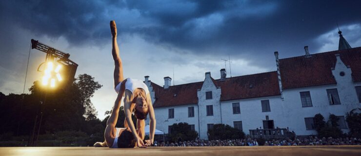 Yasmin Naghdi and Reece Clarke perform a contemporary pas de deux on an outdoor stage in front of a large manor. Naghdi bends backward with one leg extended toward the sky, her fingertips gently brushing the floor, as Reece Clarke lies on his back and supports her waist with his hands. They wear simple white costumes.