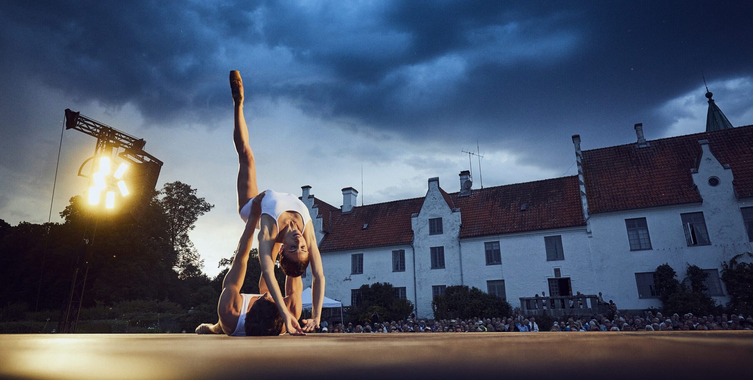 Yasmin Naghdi and Reece Clarke perform a contemporary pas de deux on an outdoor stage in front of a large manor. Naghdi bends backward with one leg extended toward the sky, her fingertips gently brushing the floor, as Reece Clarke lies on his back and supports her waist with his hands. They wear simple white costumes.