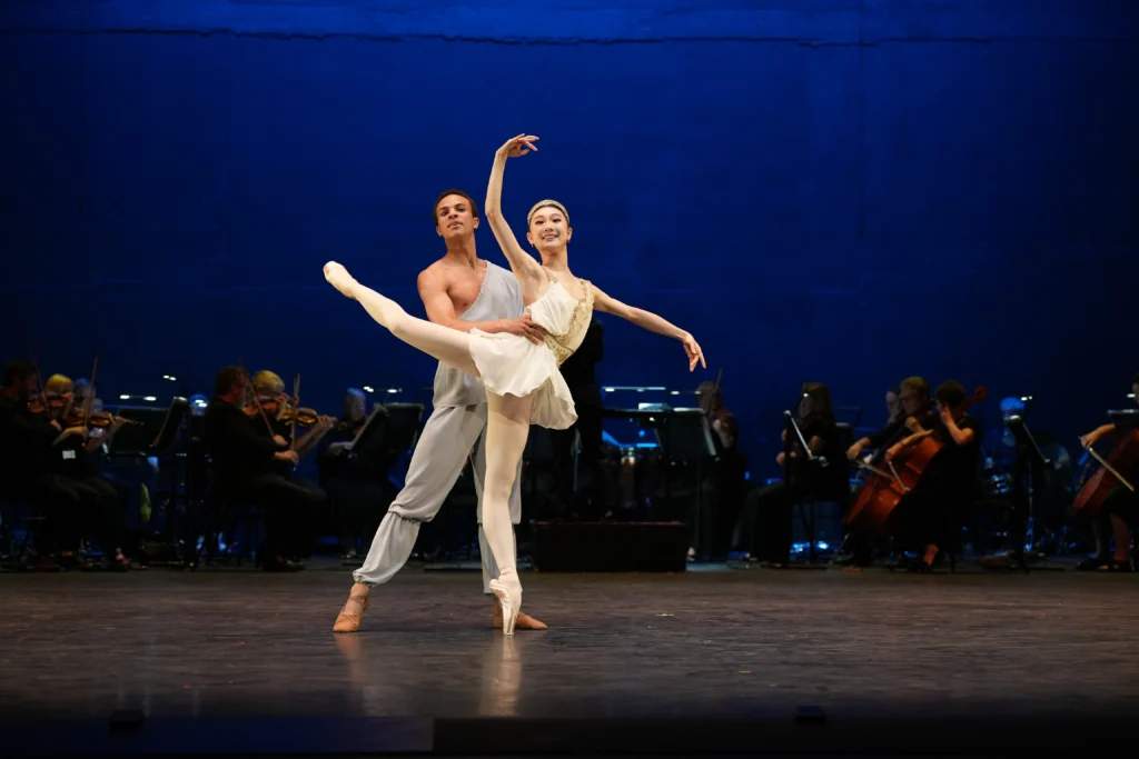 Yihan Qin and Zai Calliste perform a pas de deux onstage, Qin in a short white dress and Calliste in a light blue off-the-shoulder costume with draped fabric. They dance in front of an orchestra onstage, Calliste supporting Qin in an attitude derriere on pointe with arms in high third.