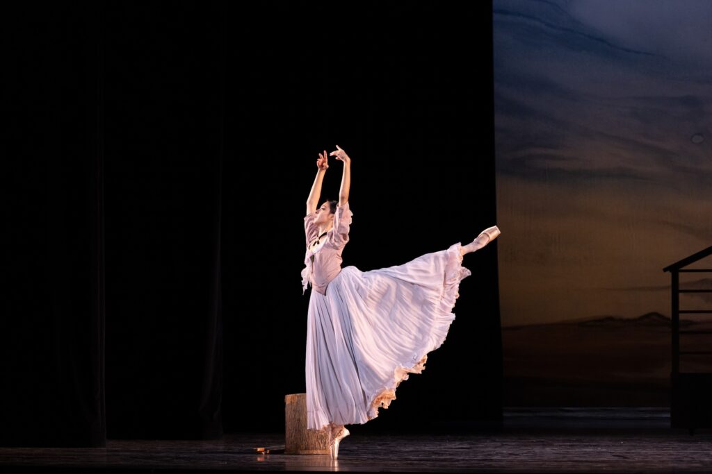 Natalie Patel, wearing a long purple prarie-style dress, does an arabesque on pointe with her arms in high fifth. She performs onstage in front of a chopping block, with dark wings and a backdrop showing a prairie setting in the background.