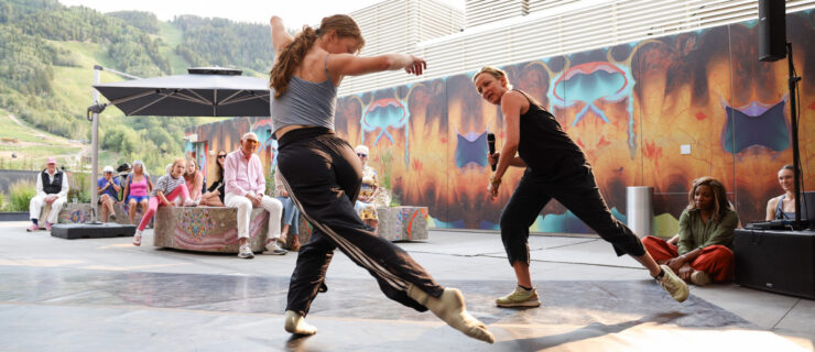 Penny Saunders leads Jane Mumford in a rehearsal demonstration at the Aspen Art Museum. They dance under a roofed outdoor pavilion as onlookers sit and watch.