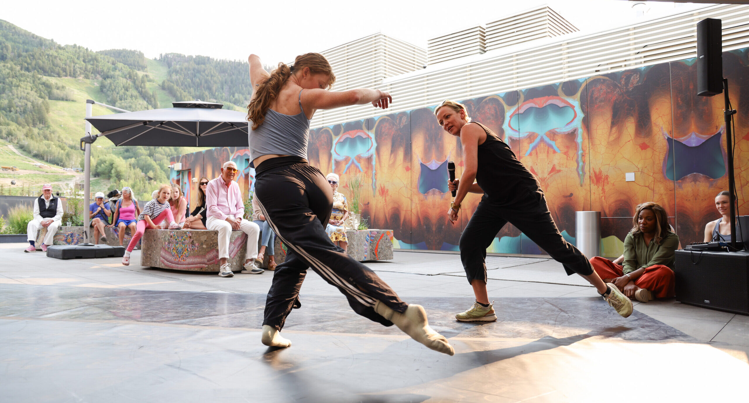 Penny Saunders leads Jane Mumford in a rehearsal demonstration at the Aspen Art Museum. They dance under a roofed outdoor pavilion as onlookers sit and watch.