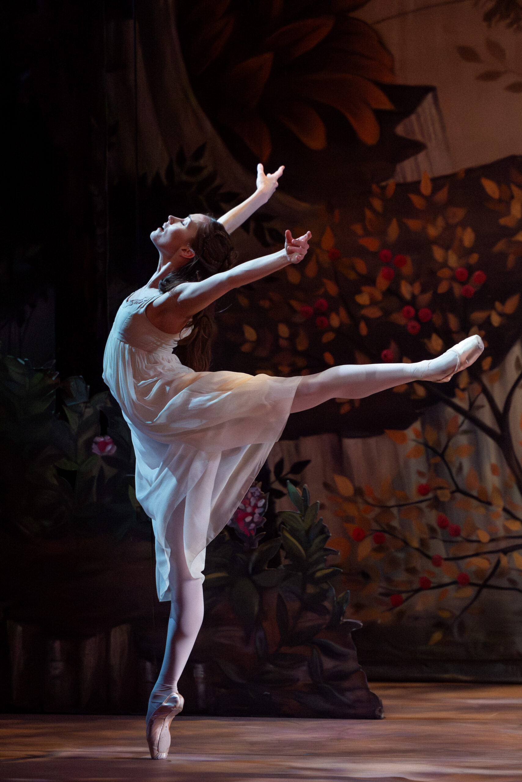Grace Carroll does an arabesque on pointe while arching back and lifting her arms in a high "V" behind her. She wears a long, white, flowing dress and closes her eyes with emotion.