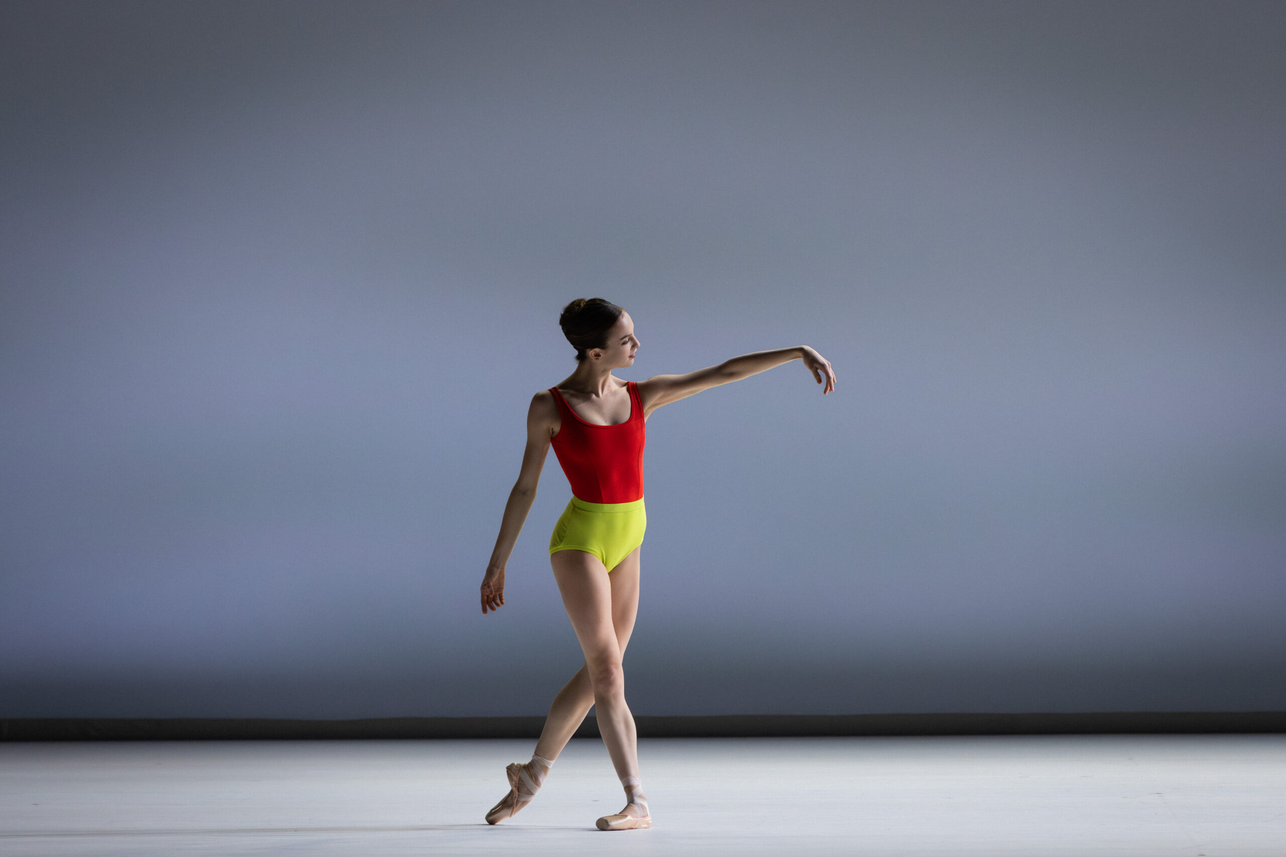 Samara Merrick stands in b-plus with one arm down and the other lifted gently to the side, her fingers curving down to the floor. She wears a sleeveless leotard with a bright red top and highlighter-yellow bottom.