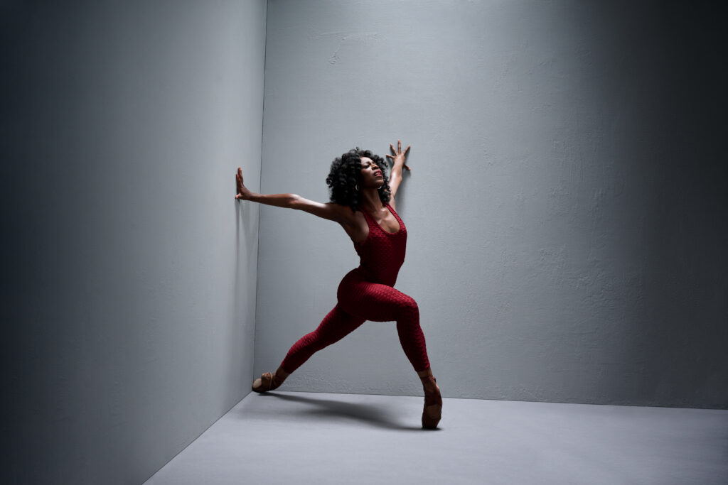 In front of a gray backdrop with two walls converging at a corner, April Watson lunges forward in her pointe shoes. She opens her chest wide so her hands rest on either wall, and her back pointe shoe reaches the corner where the walls meet the floor. She wears a dark red unitard and wears her long hair down.