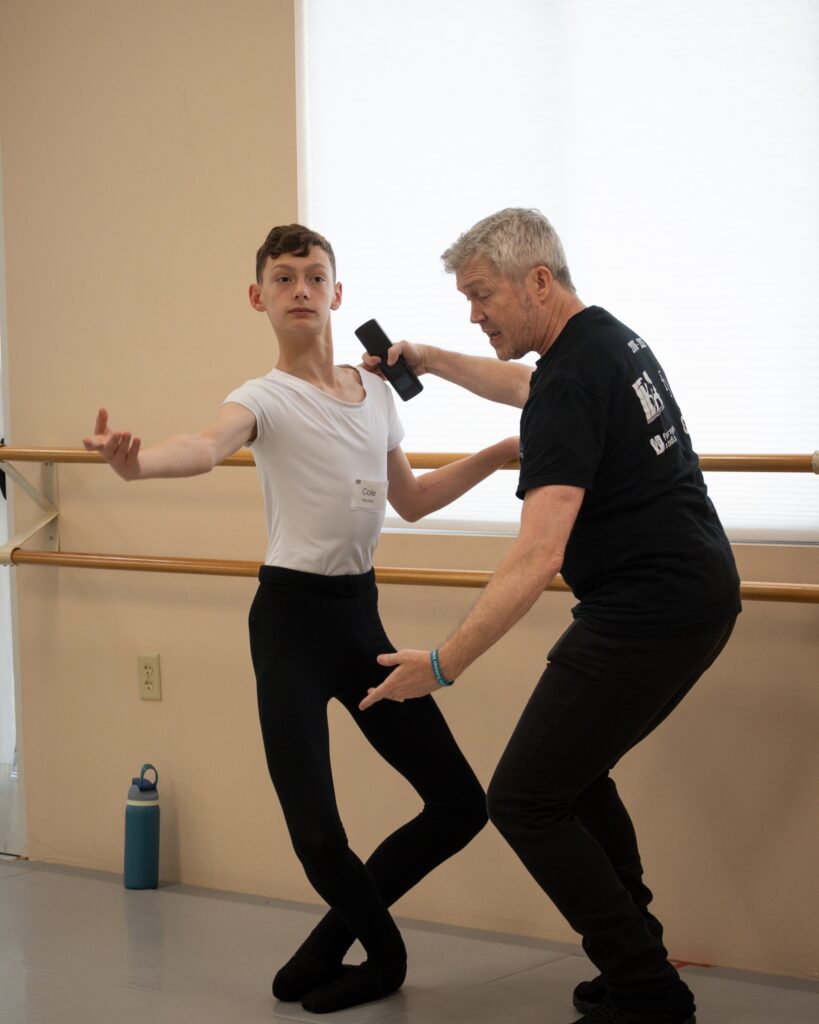 A young, teenage dancer wearing a white t-shirt, black tights and black ballet shoes does a plié in fifth position at the barre. A techer wearing a black t-shirt and pants stands directly in front of him, touching his shoulder as he coaches him through the step.