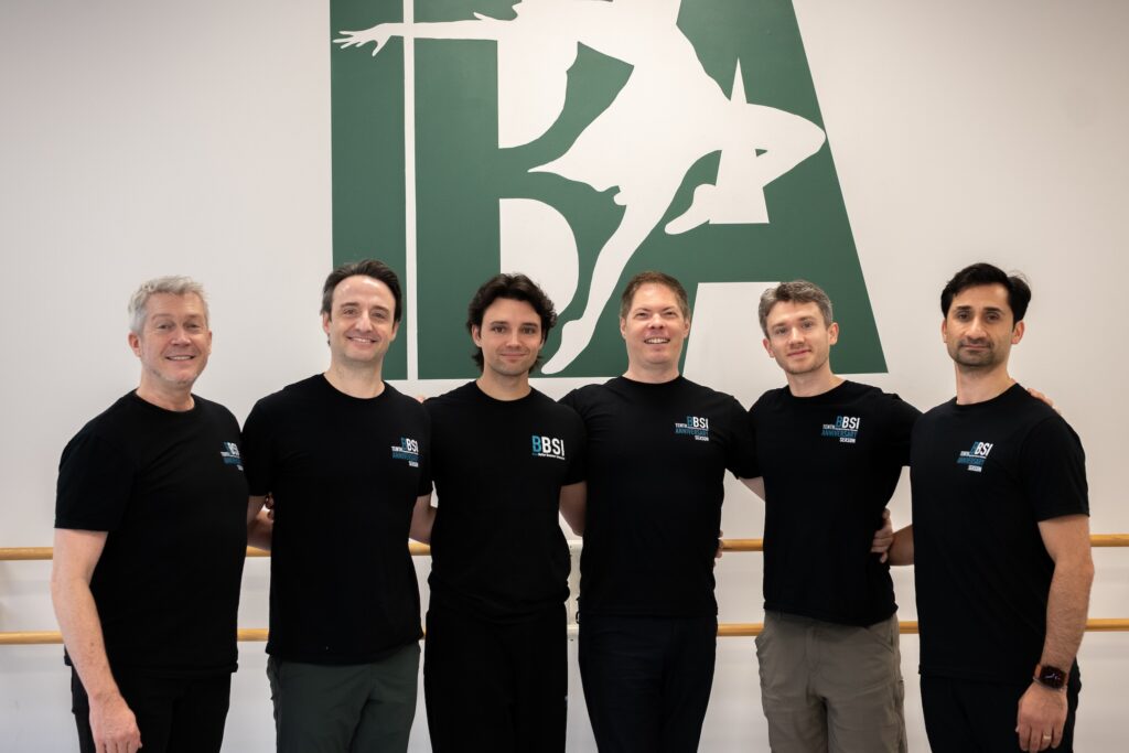 Six men wearing black t-shirts stands close together, arms around waists, and pose for a photo.