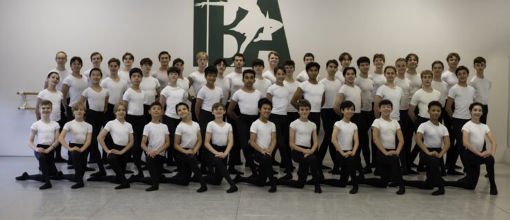 A large group of ballet studenst wearing white t-shirts and black tights and slippers pose for a photo. They pose close together in four staggered lines, with the first line kneeling on their left knee and the others standing behind them.