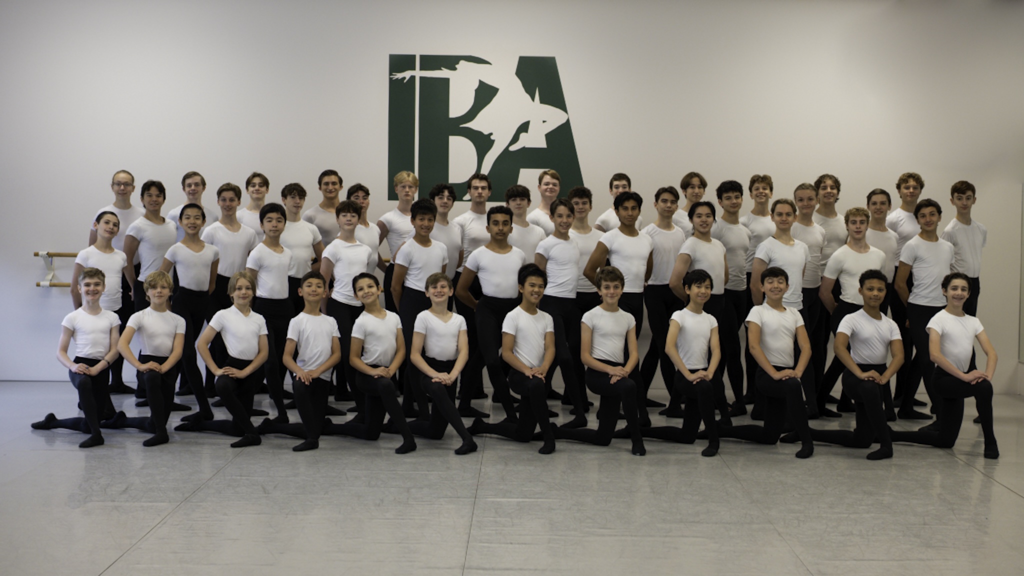 A large group of ballet students wearing white t-shirts and black tights and slippers pose for a photo. They pose close together in four staggered lines, with the first line kneeling on their left knee and the others standing behind them.