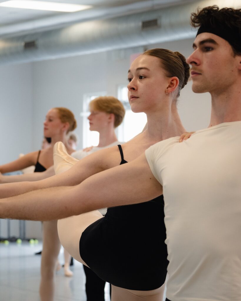 A close-up photo shows two sets of dance partners in a ballet class. The boys, wearing white ts-hirts, hold their right arms out as their partners, in black leotards, pink tights, and pointe shoes, take their extended shoulder and hand and pose in piqué attitude.