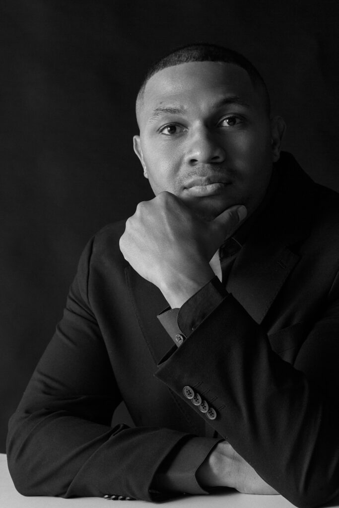 In this black and white photo, a Black man in his early 40s is shown from the waist up, wearing a sport coat with a dark shirt underneath. He rests his elbows on a table, holding his chin with his left hand as he tucks the right arm in towards his body. His gives a small, closed-mouth smile to the camera.