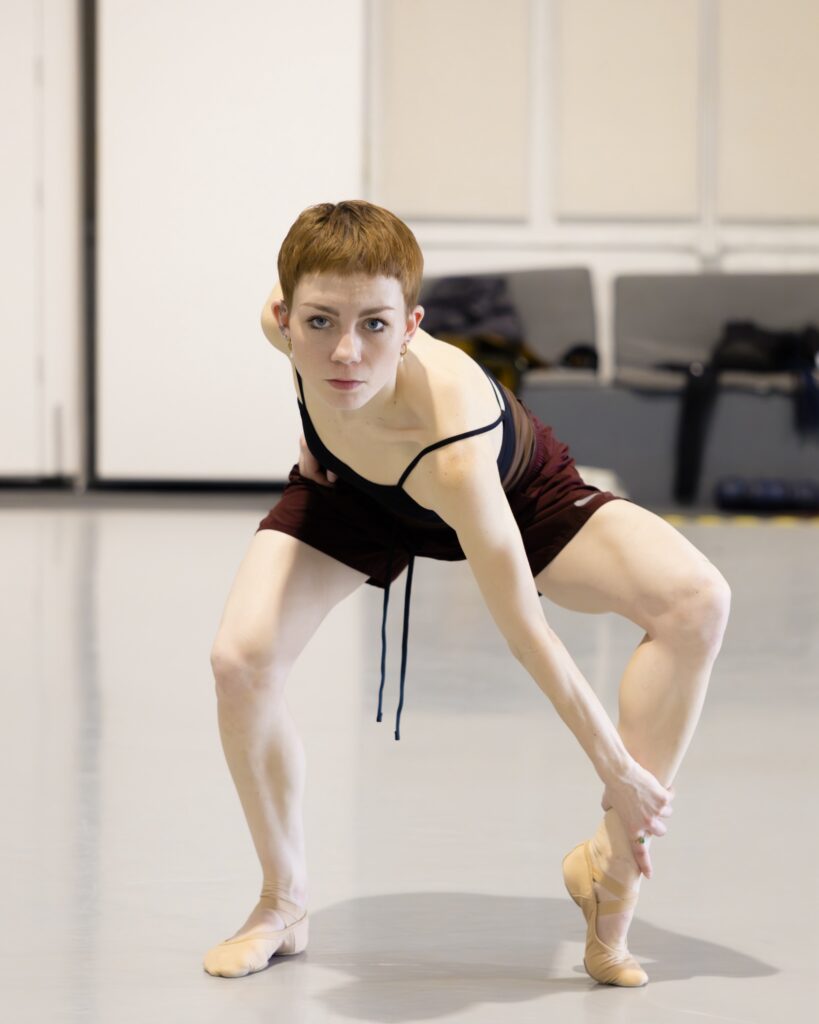 In a dance studio, Lanie Jackson, wearing a black thin-strapped leotard and burgundy shorts, crouches down slightly and grabs her left ankle with her left hand. Her right hand is on her hip and she looks straight ahead intensely.