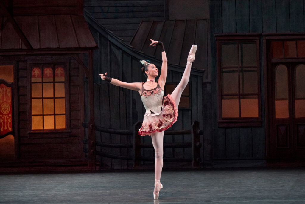 In front of a Western-inspired set piece, Brittany Pollack kicks one leg in a high battement en pointe, her arms in high third. She smiles jubilantly with a slight turn of her head. She wears a short cream, brown, and light pink dress with a ruffled skirt; brown fingerless gloves; a choker; and a feathered headpiece.
