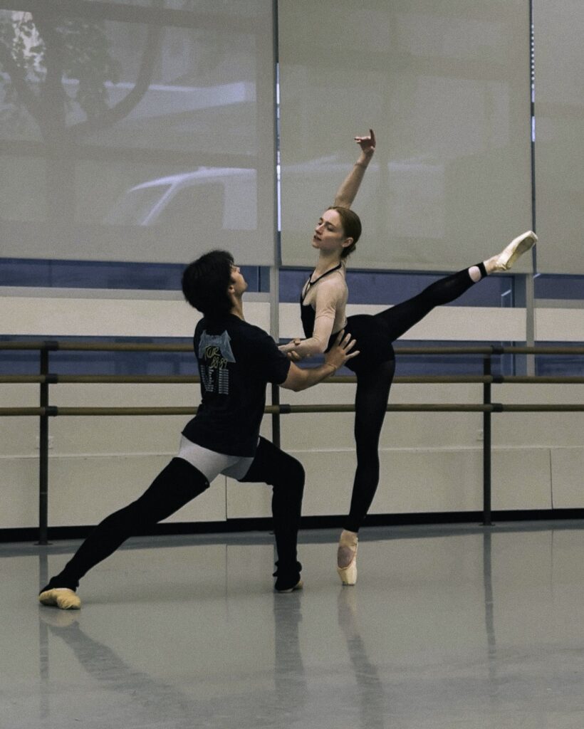 A male dancer in a black t-shirt, blue tights, and long black legwarmers faces his partner and lunges on his left leg, holding her waist as she does an arabesque towards him. The ballerina wears a black unitard and pink pointe shoes.