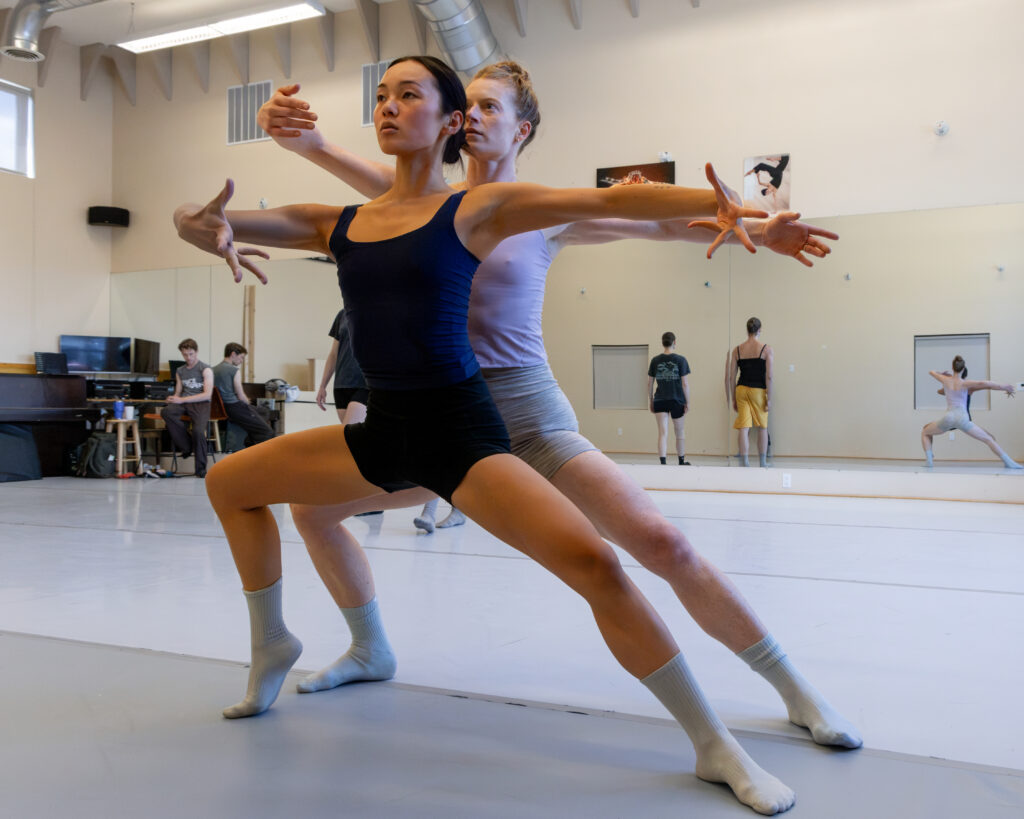 Nicole Bui stands directly in front Jane Mumford as they both lunge to the right with their left legs bent slightly. They hold their arms in a stylized third position, fingers splayed.