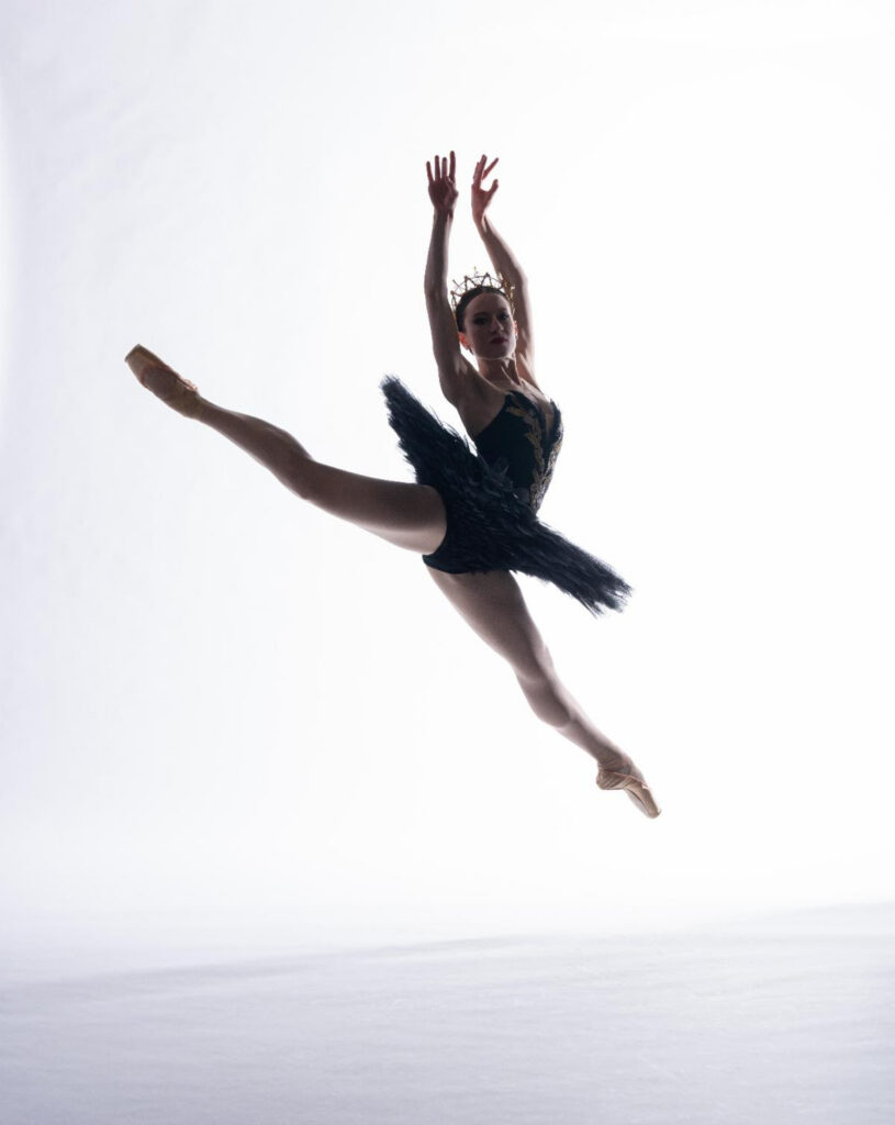 Ashley Baker, lit from behind in front of a stark white backdrop, splits her legs in the air with her back leg in a slight attitude. She lifts both arms overhead like wings and wears a black pancake tutu.