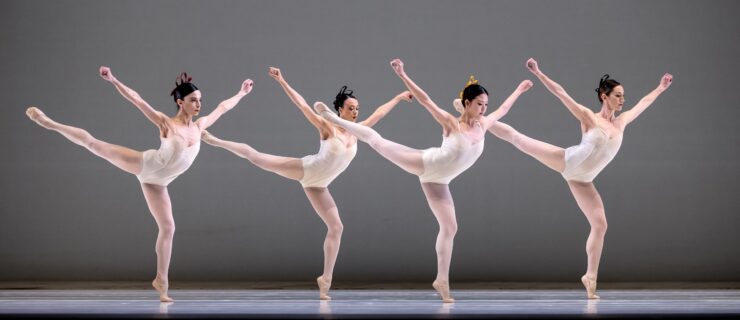 Four female dancers in white leotards, white tights, and white slippers do an arabesque facing stage left in a staggered line. They bend their supporting knee, pressing over their standing foot, and hodl their arms up in a V shape, fists clenched.