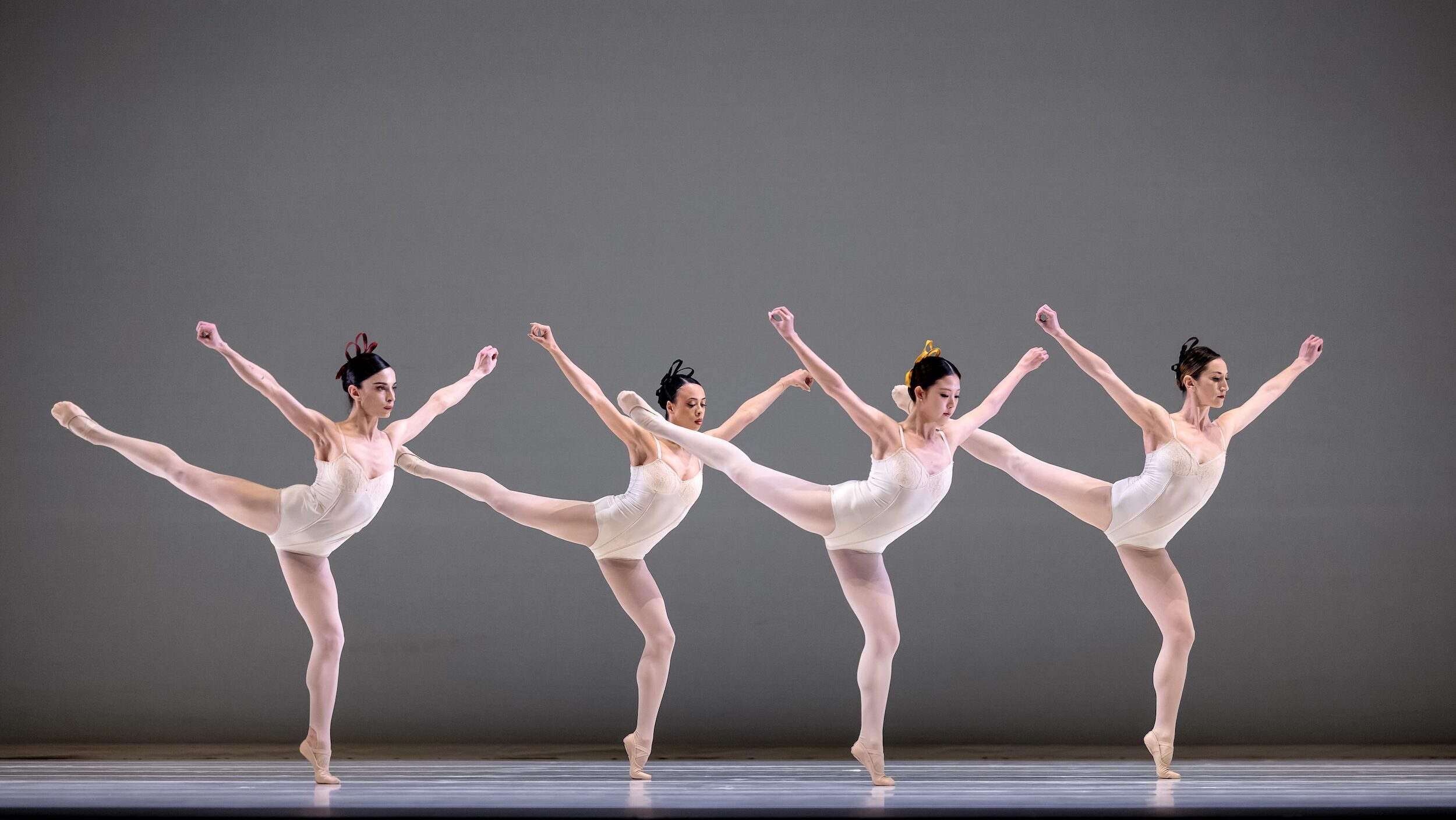 Four female dancers in white leotards, white tights, and white slippers do an arabesque facing stage left in a staggered line. They bend their supporting knee, pressing over their standing foot, and hodl their arms up in a V shape, fists clenched.