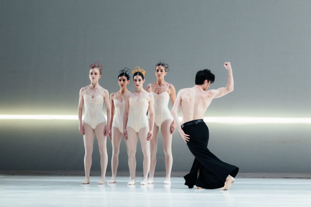 Four female dancers in white leotards, white tights, and white slippers stand close together in a staggered line. A male dancer in wide-legged black pants dances to their left, facing them, with his bent right arm raised at his side, fist closed.He looks toward his right hand and keeps his left arm down at his side as he crosses his left leg back.