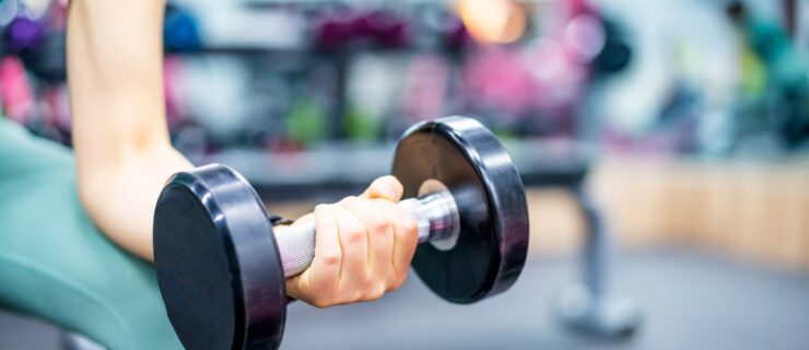 A close up photo of a woman's left arm holding a free weight against her thigh while strength training at the gym. She wears a green leggings.