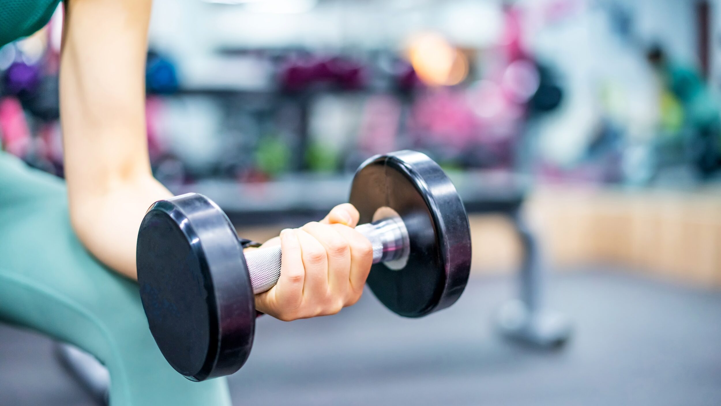 A close up photo of a woman's left arm holding a free weight against her thigh while strength training at the gym. She wears a green leggings.