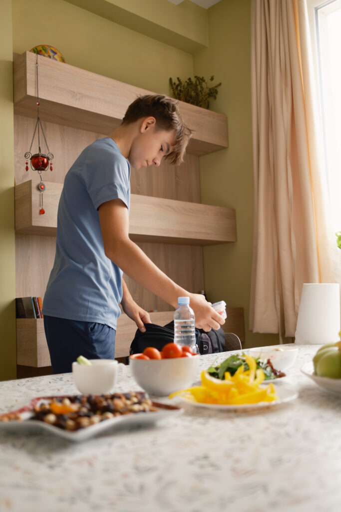 A teenage boy prepares for school by packing a backpack with lunch and other essentials in a cozy home setting. The table is filled with healthy food options.