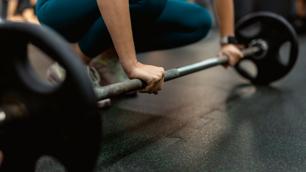 Cropped image of woman squatting down to lift a barbell at the gym. She wears turquoise leggings and sneakers.