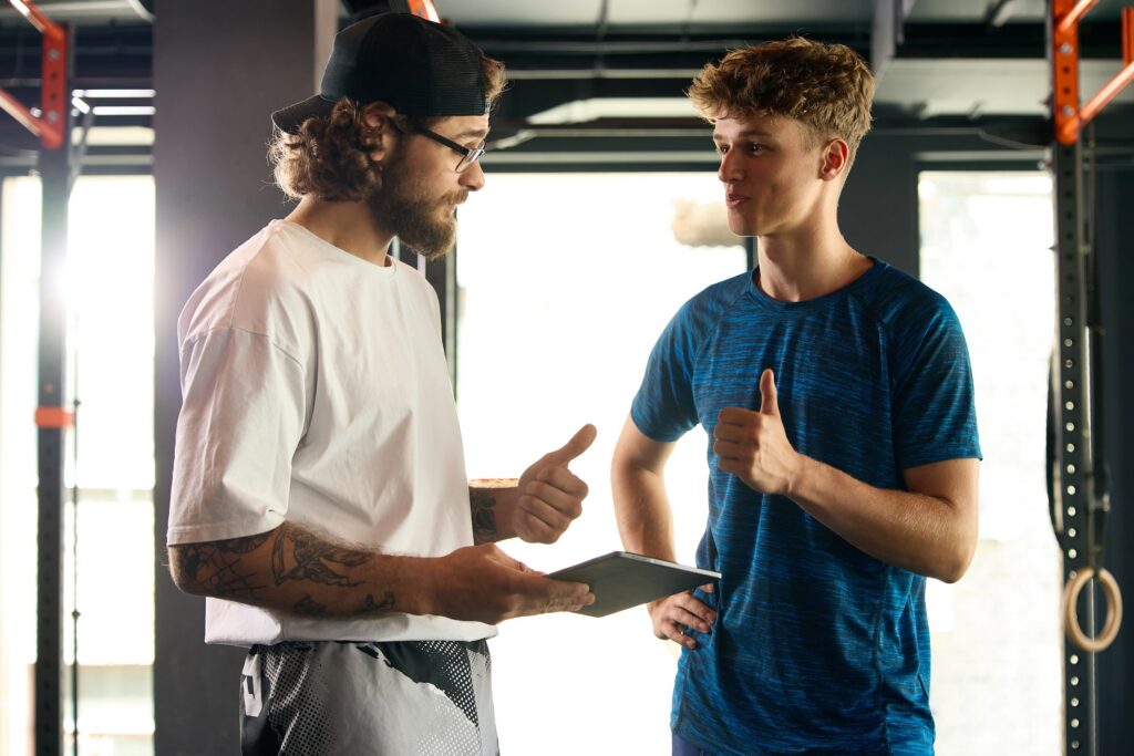 Young male dancer gives a thumbs up to personal trainer at the gym after reviewing workout results on digital tablet.
