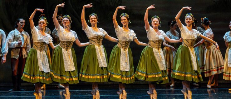 Six female dancers in peasant costumes with white blouses, green skirts, and white aprons, pose in a line facing the audience, their feet in sous-sus. They hold the waist of the woman next to them with their left hand and lift their right arm up. Other dancers mill about behind them onstage.