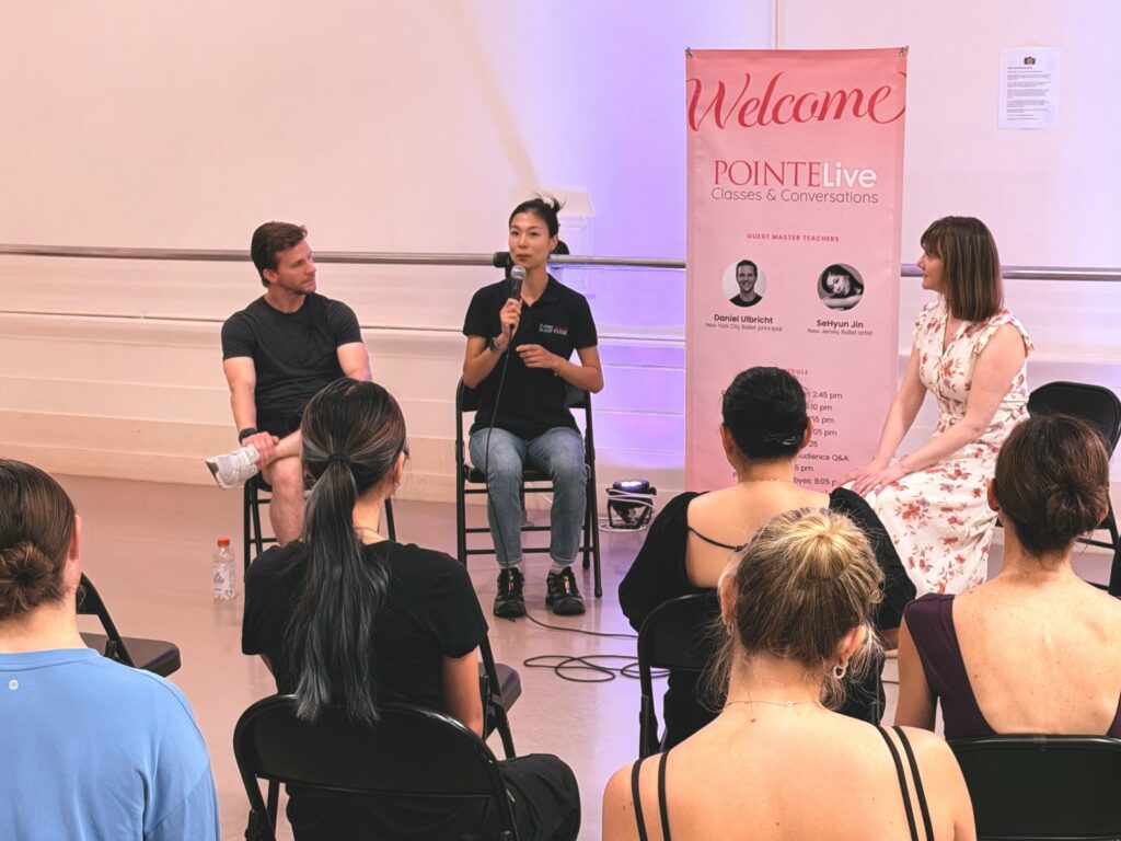 A man and two women sit in folding chairs in front of a group of adult dancers wearing practice clothes. A vertical, pink banner is placed in between the women as one speaks into a microphone.
